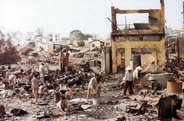 Foto van Vietnameze burgers in Cholon na het Tetoffensief. Je ziet hun kleren wassen en opruimen, ze lopen tussen het puin van kapotgeschoten huizen. Ook op de achtergrond zie je meerdere gebouwen die gedeeltelijk ingestort zijn.