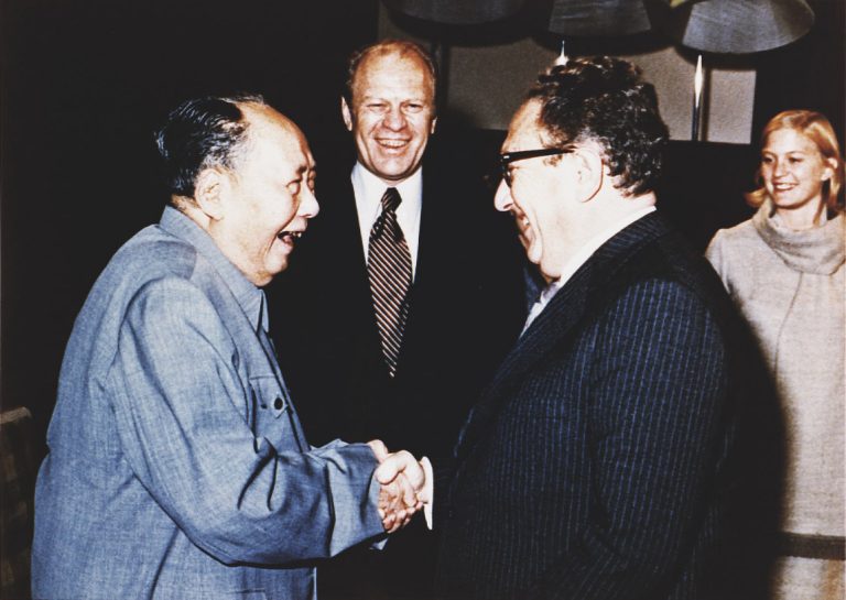 Foto van een lachende Kissinger die de hand van een lachende Mao Zedong schudt, op de achtergrond zie je Gerald Ford lachen.